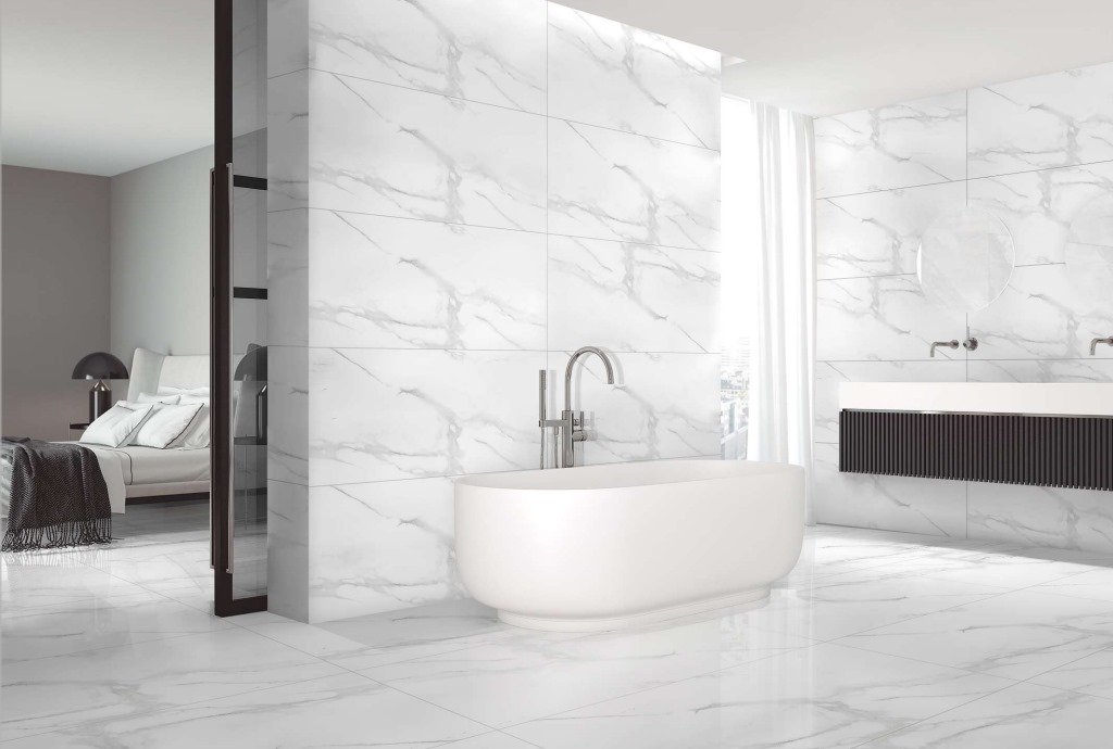 Modern bathroom with white marble walls and floor, a freestanding oval bathtub, wall-mounted faucet, and a sleek floating vanity with round mirrors. A bedroom with a bed and side table is visible through the glass door.